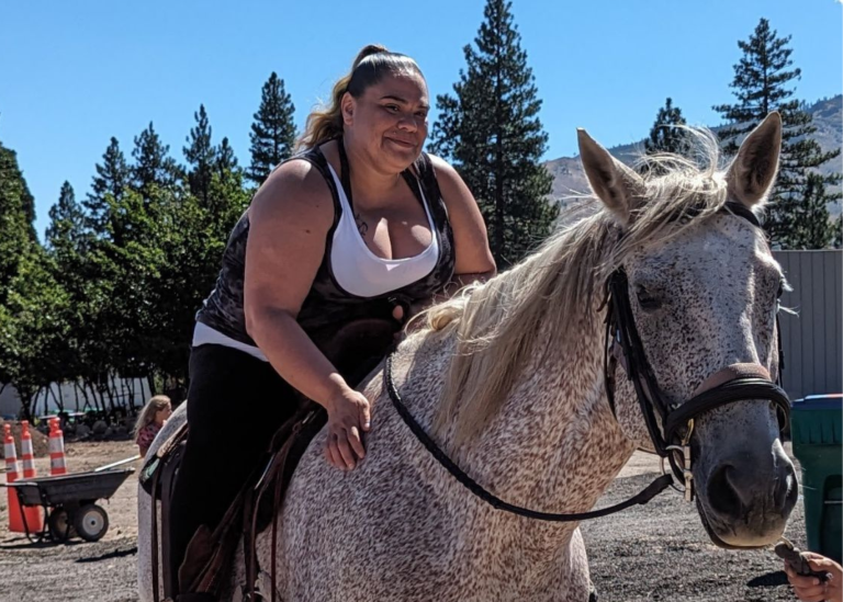 empowerment center client Nicole Hulihee riding a horse with pine trees and grassy nevada hills in background